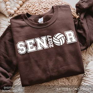 Op de afbeelding: Een donkerbruin sweatshirt met ronde hals en witte blokletters met de tekst SENIOR en het jaartal 2027. Een witte volleybalafbeelding vervangt de letter O in het woord SENIOR.