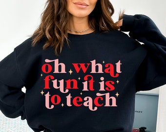 May include: Black sweatshirt with the phrase "Oh what fun it is to teach" in red and pink lettering. The text is accented with small gold stars. A casual, comfortable top for teachers.