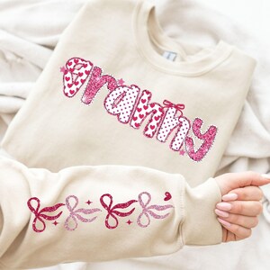 May include: A beige sweatshirt with the word "Granny" in pink glitter lettering, with a heart and polka dot pattern. The sleeves feature pink glitter bows and stars. A cozy and stylish gift for grandmothers.