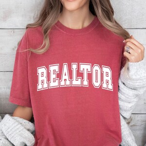 May include: A red t-shirt with the word "REALTOR" printed in white on the front. The shirt has a vintage, distressed look.