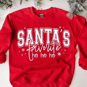 May include: A red sweatshirt with the text "SANTA'S favorite ho ho ho" in white. The text is in a collegiate style font. The sweatshirt has long sleeves and a crew neck.