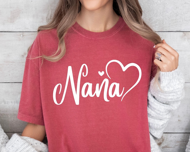 Puede incluir: Una camiseta roja con la palabra "Nana" en letras blancas cursivas y un dise&ntilde;o de coraz&oacute;n blanco.