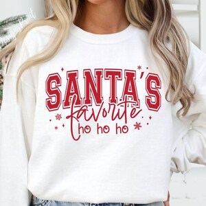 May include: White sweatshirt with the text "SANTA'S favorite ho ho ho" in red. The text is in a collegiate style font with a red outline. The design includes snowflakes and stars. The sweatshirt is a festive holiday item.