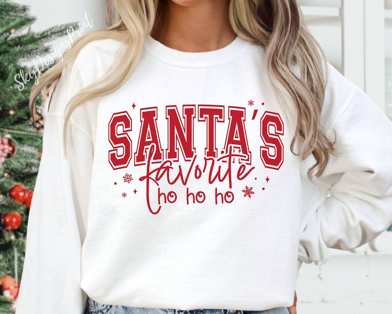 May include: White sweatshirt with the text "SANTA'S favorite ho ho ho" in red. The text is in a collegiate style font with a red outline. The design includes snowflakes and stars. The sweatshirt is a festive holiday item.