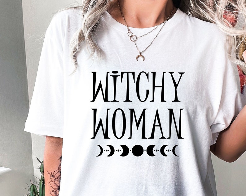 May include: White t-shirt with black text that reads "Witchy Woman" and a graphic of five crescent moons.
