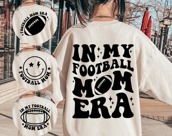 May include: White sweatshirt with black text that reads "In My Football Mom Era" with a football graphic. The text is in a retro font style.