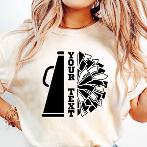 Pu&ograve; includere: T-shirt color crema con grafica in bianco e nero di un megafono e un pompon. Il testo "YOUR TEXT" &egrave; allineato verticalmente a sinistra del design del pompon. La maglietta &egrave; realizzata in materiale morbido.