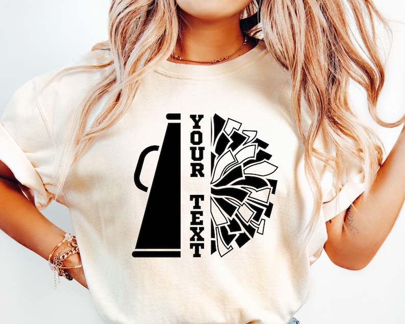 Pu&ograve; includere: T-shirt color crema con grafica in bianco e nero di un megafono e un pompon. Il testo "YOUR TEXT" &egrave; allineato verticalmente a sinistra del design del pompon. La maglietta &egrave; realizzata in materiale morbido.