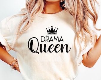 May include: Cream-colored t-shirt with the words "DRAMA Queen" in black script, topped with a black crown graphic. The shirt is a casual style, likely made of soft fabric, and suitable for everyday wear. The design is centered on the chest.