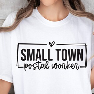 Op de afbeelding: Wit T-shirt met zwarte tekst die "Small Town -postal worker-" in een kader met een hart boven de tekst luidt.
