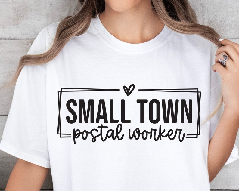 Op de afbeelding: Wit T-shirt met zwarte tekst die "Small Town -postal worker-" in een kader met een hart boven de tekst luidt.