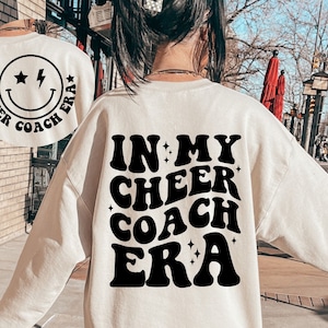May include: A white sweatshirt with black text that reads "In My Cheer Coach Era" in a retro font. The text is surrounded by small stars.