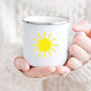 May include: A white enamel mug with a yellow sun graphic. The mug is being held by a person wearing a white sweater.