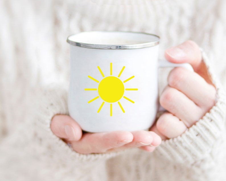 May include: A white enamel mug with a yellow sun graphic. The mug is being held by a person wearing a white sweater.