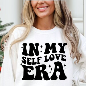 May include: White sweatshirt with black text that reads "In My Self Love Era" in a retro font.