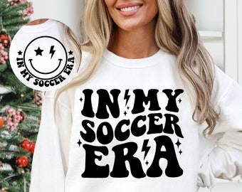 Może przedstawiać: Biała bluza z czarnym napisem "In My Soccer Era" w retro czcionce. Tekst jest otoczony małymi gwiazdkami i błyskawicami.