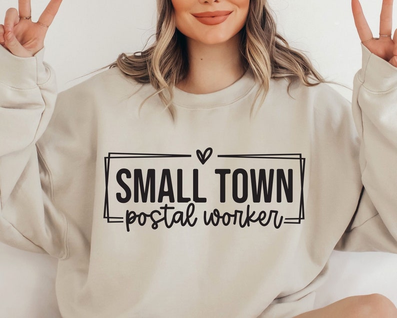 Op de afbeelding: Een beige sweatshirt met de tekst "SMALL TOWN postal worker" in zwarte letters in een zwart kader met een klein hartje boven de tekst.