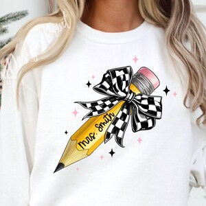 Puede incluir: Sudadera blanca con un l&aacute;piz amarillo atado con un lazo a cuadros blanco y negro. El l&aacute;piz tiene el texto "Mrs. Smith" escrito en &eacute;l.