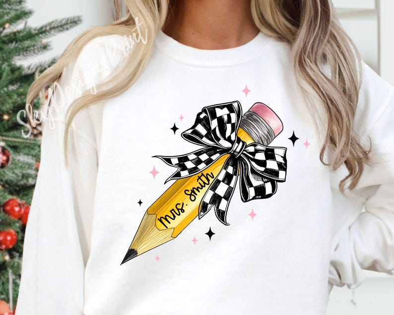Puede incluir: Sudadera blanca con un l&aacute;piz amarillo atado con un lazo a cuadros blanco y negro. El l&aacute;piz tiene el texto "Mrs. Smith" escrito en &eacute;l.