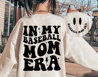 Può includere: Una felpa bianca con la scritta "IN MY BASEBALL MOM ERA" stampata in nero sul retro. La parte anteriore della felpa ha un viso sorridente con la scritta "BASEBALL MOM ERA" intorno.
