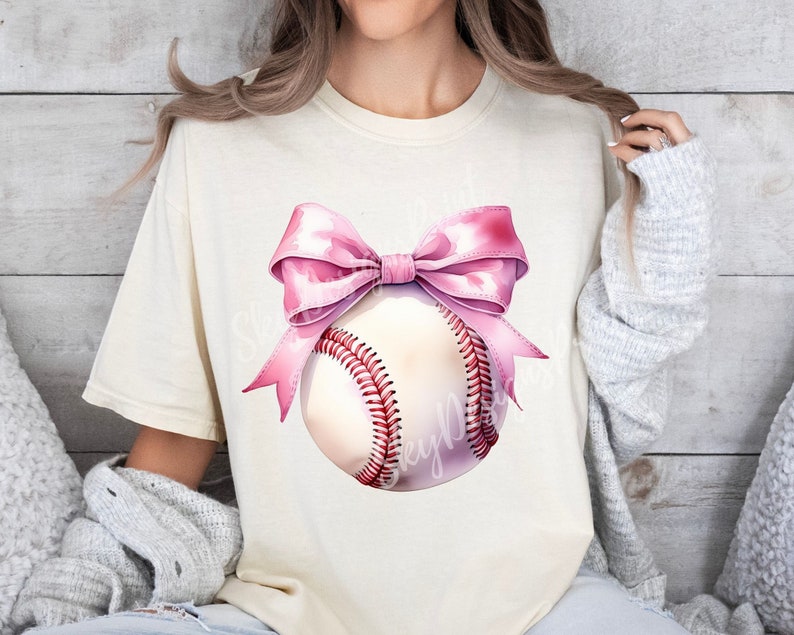 May include: A white baseball with a pink bow on top. The baseball has red stitching and is on a white t-shirt.