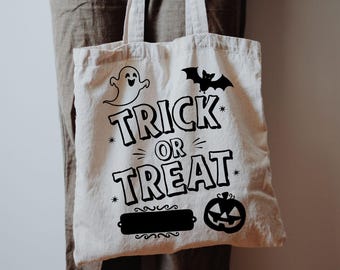 May include: A cream-colored tote bag with black Halloween-themed graphics. The bag features a ghost, bat, and jack-o'-lantern, with the words "TRICK OR TREAT" in bold letters. The bag has two handles and is made of a sturdy fabric.