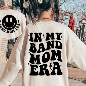 Pu&ograve; includere: Felpa bianca con testo nero che recita "In My Band Mom Era" in un font retr&ograve;. La parte posteriore della felpa ha una faccina sorridente nera con il testo "Band Mom Era" intorno.