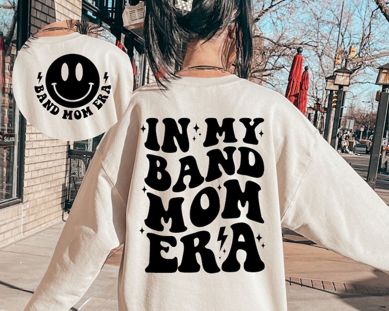Pu&ograve; includere: Felpa bianca con testo nero che recita "In My Band Mom Era" in un font retr&ograve;. La parte posteriore della felpa ha una faccina sorridente nera con il testo "Band Mom Era" intorno.