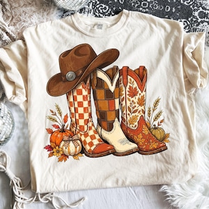 Puede incluir: Una camiseta de color crema con un estampado gráfico de un sombrero de vaquero marrón sobre tres botas de vaquero. Las botas muestran patrones de cuadros y hojas de otoño. El diseño incluye calabazas naranjas, trigo y follaje otoñal.
