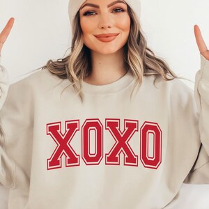 May include: Beige sweatshirt with the word "XOXO" in bold red letters. The letters have a white outline. The sweatshirt is a neutral color and has a crew neck. The image shows a person wearing the sweatshirt.