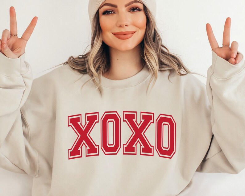 May include: Beige sweatshirt with the word "XOXO" in bold red letters. The letters have a white outline. The sweatshirt is a neutral color and has a crew neck. The image shows a person wearing the sweatshirt.