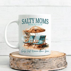 May include: White ceramic mug with a blue and white illustration of beach chairs under a beach umbrella. The text "Salty Moms Beach Club" and "Girls Just Wanna Have Sun" are printed on the mug.
