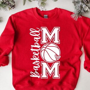 Może przedstawiać: Czerwona bluza z białym napisem "Basketball Mom" z grafiką koszyk&oacute;wki i serca.