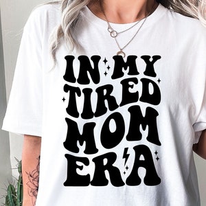 以下が含まれることがあります： レトロなフォントスタイルで「In My Tired Mom Era」と書かれた黒いテキストがプリントされた白いTシャツ。テキストは小さな星と稲妻で飾られています。