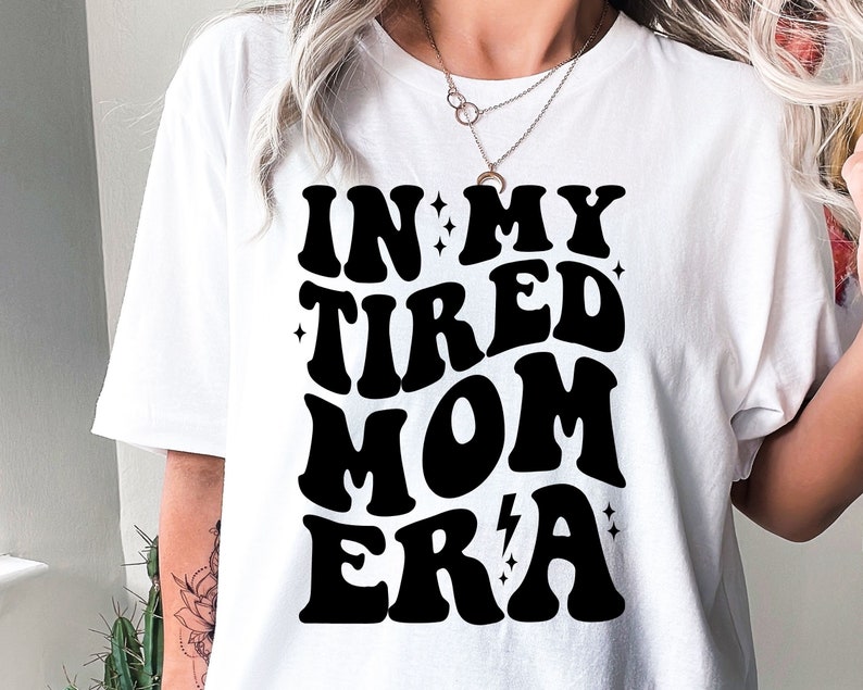 以下が含まれることがあります： レトロなフォントスタイルで「In My Tired Mom Era」と書かれた黒いテキストがプリントされた白いTシャツ。テキストは小さな星と稲妻で飾られています。