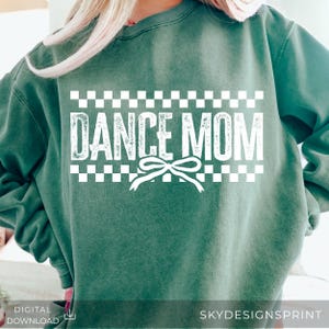 Könnte beinhalten: Ein waldgrünes Sweatshirt mit Rundhalsausschnitt und weißem Grafikdruck. Das Design umfasst einen Schachbrettmuster-Rahmen über und unter dem Text DANCE MOM sowie ein kleines Schleifensymbol unter dem Wort DANCE. Der Druck hat eine abgenutzte Textur.