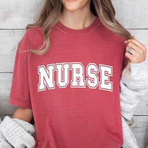 May include: A red t-shirt with the word "NURSE" printed in white letters.