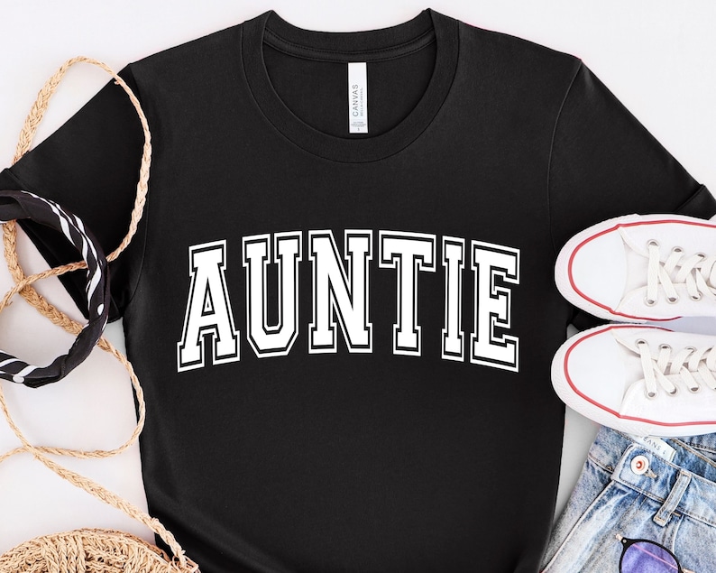 May include: Black t-shirt with white text that reads "AUNTIE" in a varsity style font.