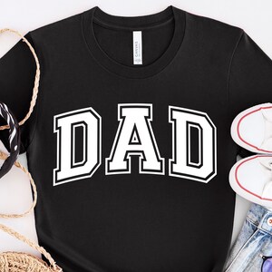Op de afbeelding: Zwart T-shirt met witte tekst "DAD" in een varsity-letter-stijl.