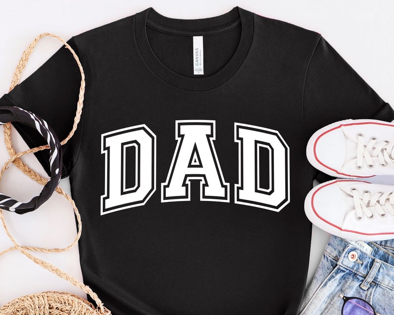 Op de afbeelding: Zwart T-shirt met witte tekst "DAD" in een varsity-letter-stijl.