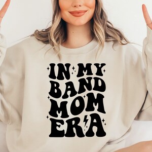 Pu&ograve; includere: Una felpa color crema con una grafica nera che recita "In My Band Mom Era" in un font retr&ograve;. La grafica include stelle e un fulmine.