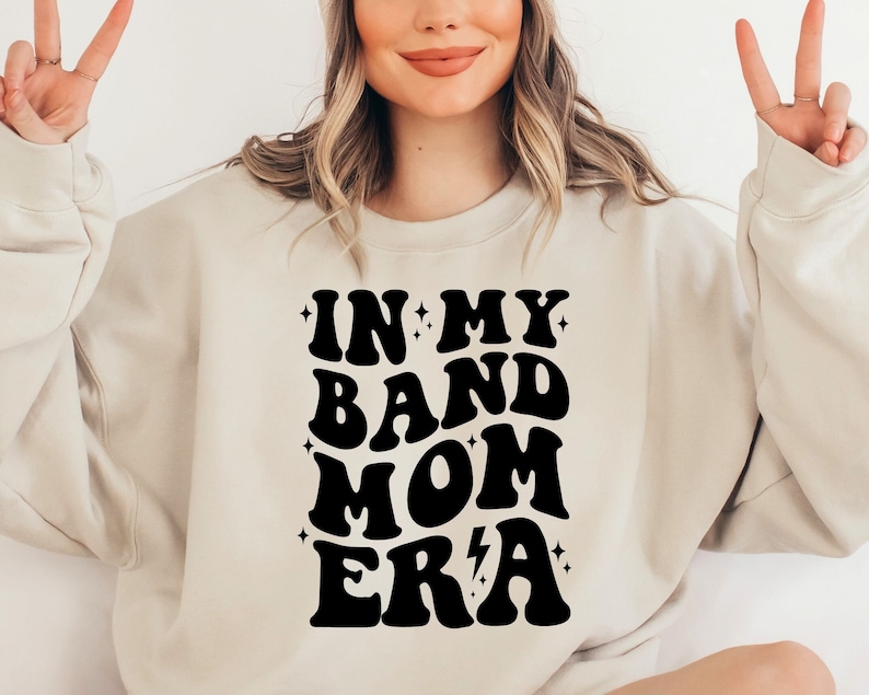 Pu&ograve; includere: Una felpa color crema con una grafica nera che recita "In My Band Mom Era" in un font retr&ograve;. La grafica include stelle e un fulmine.