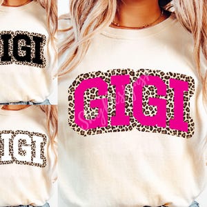 May include: Cream-colored t-shirts with the word "GIGI" in a bold, block letter style. The letters are in black, white, and pink, with a leopard print border. The shirts are casual wear.
