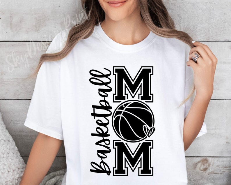 Może przedstawiać: Biała koszulka z czarnym napisem "Basketball Mom" z grafiką koszyk&oacute;wki i serca.