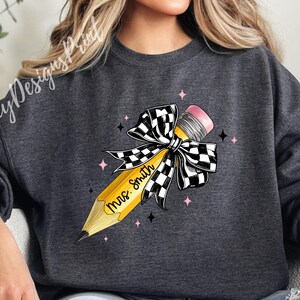 Puede incluir: Una sudadera gris oscuro con un dise&ntilde;o de l&aacute;piz amarillo. El l&aacute;piz est&aacute; atado con un lazo a cuadros blanco y negro y tiene el texto "Mrs. Smith" escrito en &eacute;l.