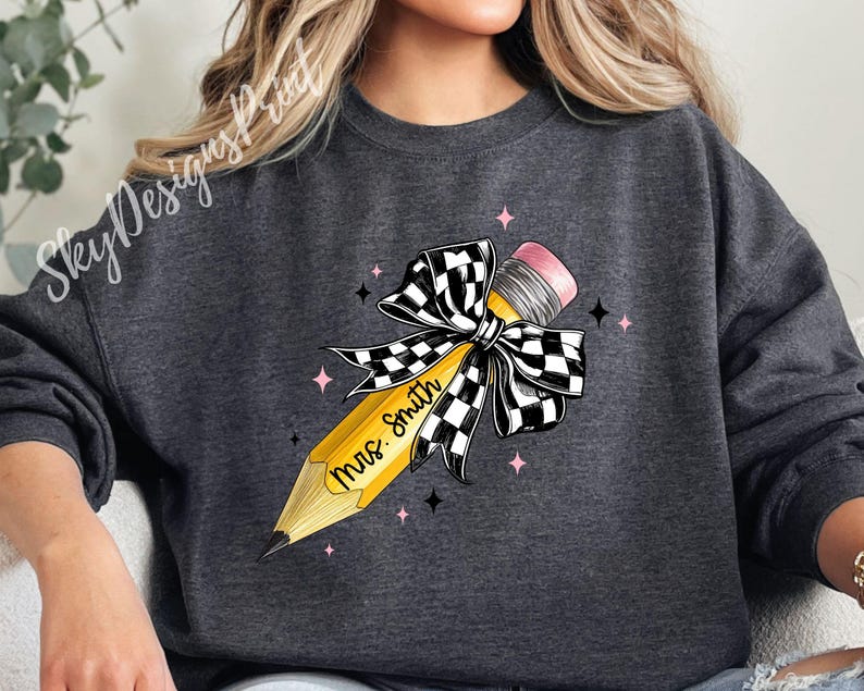 Puede incluir: Una sudadera gris oscuro con un dise&ntilde;o de l&aacute;piz amarillo. El l&aacute;piz est&aacute; atado con un lazo a cuadros blanco y negro y tiene el texto "Mrs. Smith" escrito en &eacute;l.