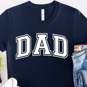 Op de afbeelding: Een marineblauw t-shirt met het woord "DAD" in witte blokletters. De letters hebben een witte omtrek en zijn in een varsity-stijl lettertype.
