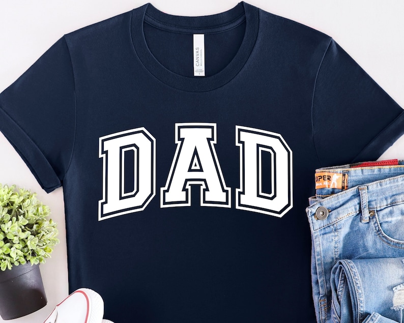 Op de afbeelding: Een marineblauw t-shirt met het woord "DAD" in witte blokletters. De letters hebben een witte omtrek en zijn in een varsity-stijl lettertype.