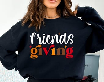 May include: Black sweatshirt with the words "friends giving" in white, orange, brown, and red lettering. The word "friends" is in a script font, and the word "giving" is in a bold, block font. The sweatshirt has a crew neck.