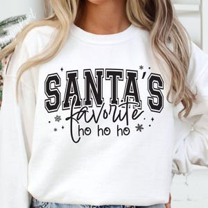 May include: White sweatshirt with the text "SANTA'S FAVORITE" in black block letters, with "ho ho ho" below. The design includes snowflakes and stars. A festive holiday-themed top.